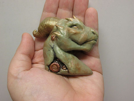 A magnet in the shape of a dragon's head, embellished with gear shapes at the neck and dusting of a bronzey mica powder pigment.