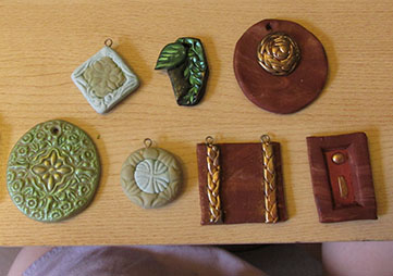 Various clay trinkets: a round green medallion with an embossed pattern, dusted with glittery gold mica powder pigment; two small clay pendants made with a cutout-and-replacement of contrasting color and then stamped; a black hexagon with a leaf pattern colored in green mica pigment powder, and a similarly-colored green leaf affixed; a square brown pendant with gold-tone braids affixed; a round brown medallion pendant with a curled gold-tone braid affixed; a pendant consisting of a brown frame opening onto a red 'canvas' colored with mica powder pigment, and abstract gold-tone shapes affixed.